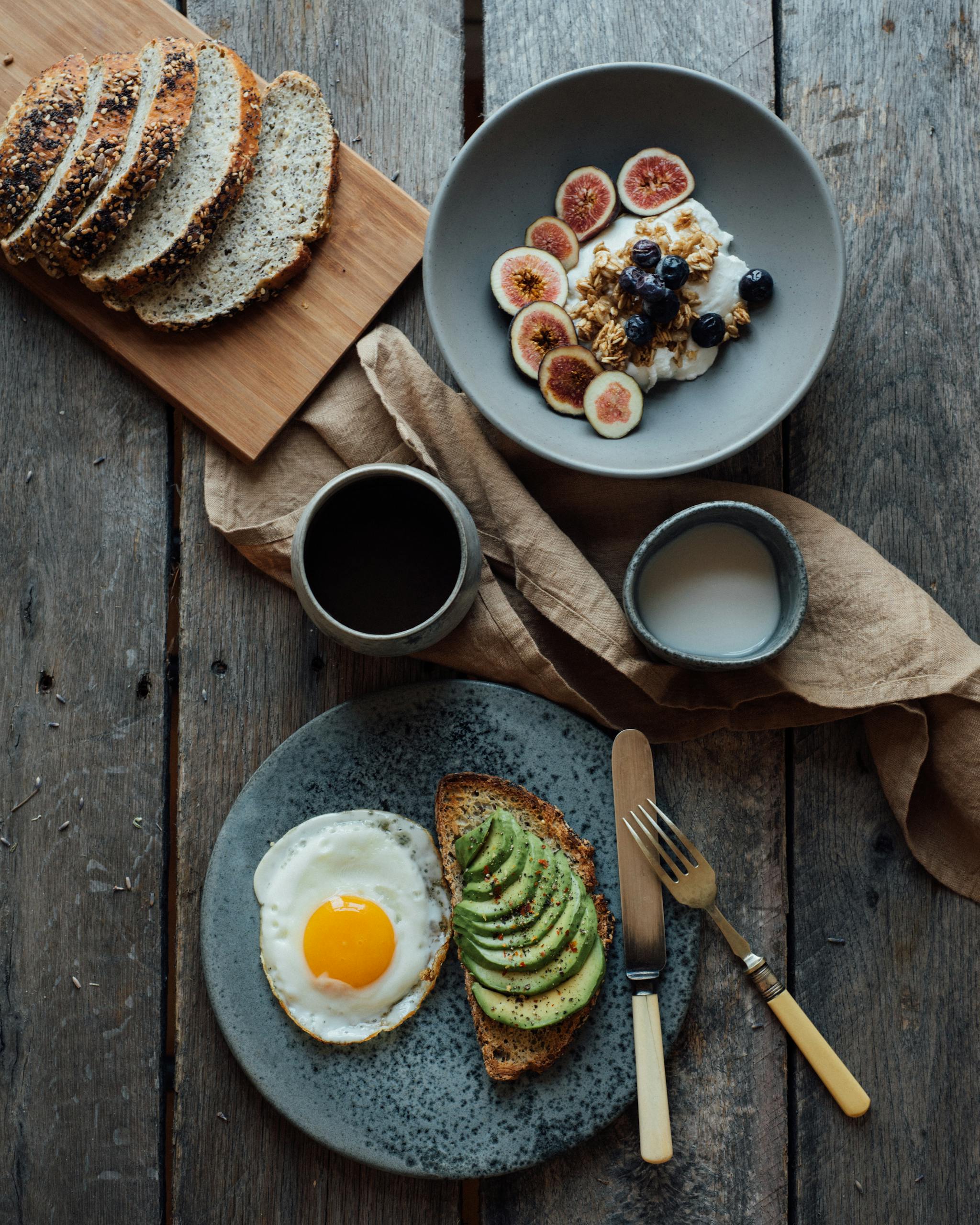 Healthy breakfast with avocado toast, eggs, and yogurt with figs and berries on a wooden table.